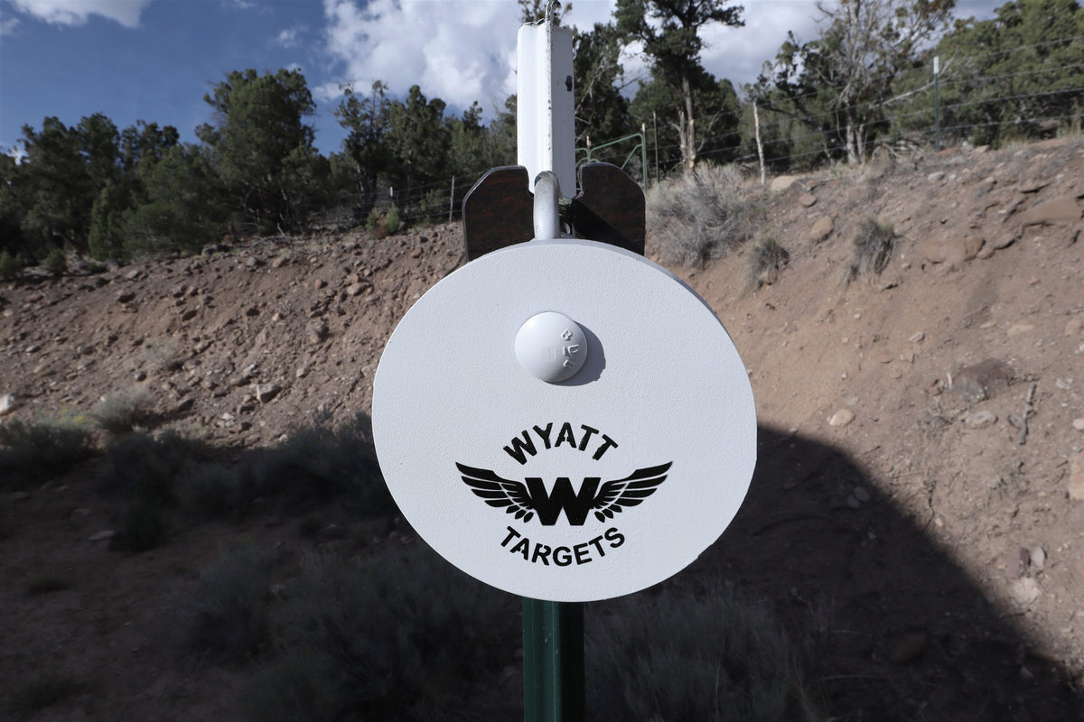Real photo: white Wyatt AR500 gong with the Wyatt Targets logo hanging from a Wyatt T-post hanger on a green T-post at the range